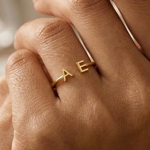 Könnte beinhalten: Ein goldfarbener Ring mit den Buchstaben "A" und "E" wird an einem Finger gezeigt. Der Ring ist zierlich und dünn, wobei die Buchstaben oben auf dem Finger positioniert sind. Der Hintergrund ist weich verschwommen, wodurch der Ring hervorgehoben wird.