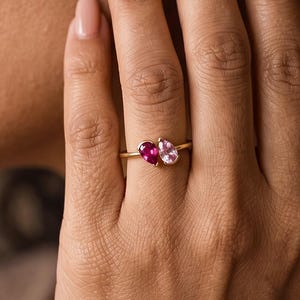 Peut inclure: Une bague en or ornée de deux pierres précieuses en forme de poire, disposées côte à côte sur un anneau fin. L'une des pierres est d'un rouge profond et vibrant, tandis que l'autre est d'un rose pâle. La bague est portée sur un doigt.