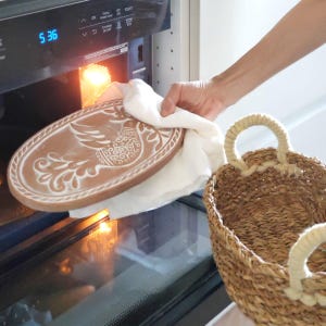 May include: An oval, brown ceramic serving platter with a white bird design is being removed from an oven. The platter is held with a white cloth. A woven basket with rope handles is in the foreground.