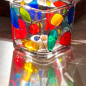 May include: A clear glass votive candle holder with a square shape. The holder is decorated with colorful, abstract shapes in red, blue, yellow, and green, outlined in black. A lit white candle is inside, casting colorful reflections.