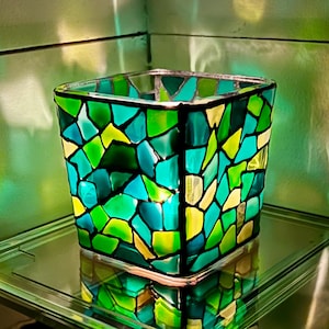 May include: Square glass candle holder with a mosaic design. The mosaic features irregular shapes in shades of green, teal, and yellow. The candle holder is sitting on a glass shelf.