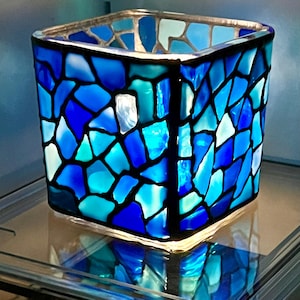 May include: Square glass candle holder with a mosaic design in shades of blue and teal. The glass pieces are outlined in black, creating a stained-glass effect. The candle holder is illuminated from within, casting a soft glow.