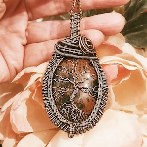 Op de afbeelding: Een hanger van koperdraad met een bruine en zwarte boom des levens steen. De hanger is ovaalvormig en heeft een koperen draadbeugel.