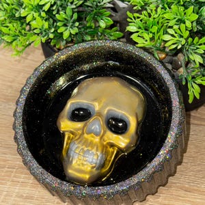 Könnte beinhalten: Eine schwarze und goldene Glitzer-Harzschale mit einem goldenen Schädel im Inneren. Der Schädel hat schwarze Augen und ein detailliertes Design.
