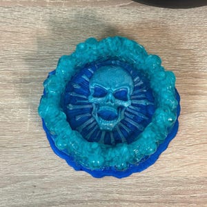 Könnte beinhalten: Eine tealblaue Harzkranzschale mit einem Schädelmotiv in der Mitte. Der Schädel ist von einem Ring aus Knochen umgeben.