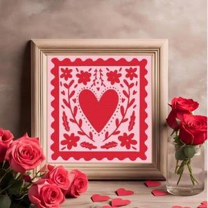 Puede incluir: Una impresión enmarcada con un fondo rosa y un corazón rojo rodeado de flores y hojas. El marco es de madera marrón claro y la impresión tiene un borde festoneado.