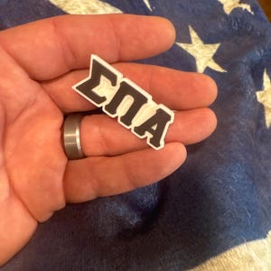 May include: A white and black 3D printed pin with the letters "&Sigma;&Pi;&Alpha;" in black.