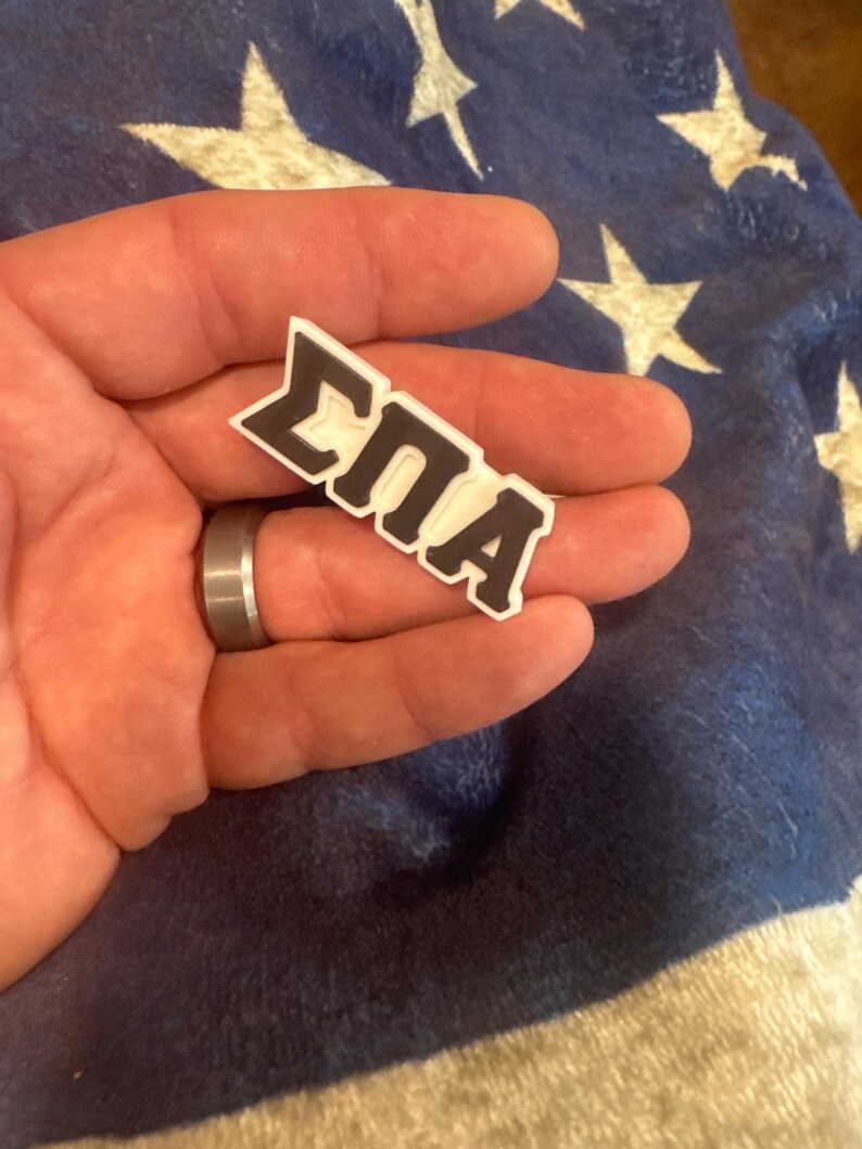 May include: A white and black 3D printed pin with the letters "&Sigma;&Pi;&Alpha;" in black.