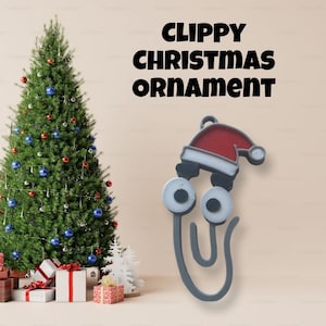 May include: A gray paperclip Christmas ornament with a red Santa hat and googly eyes. The ornament is hanging from a string and is in front of a Christmas tree.