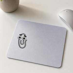 May include: A light gray mousepad featuring a cartoon paperclip character with googly eyes and headphones. The paperclip is gray with black accents. A small potted plant and a silver computer mouse are in the background.