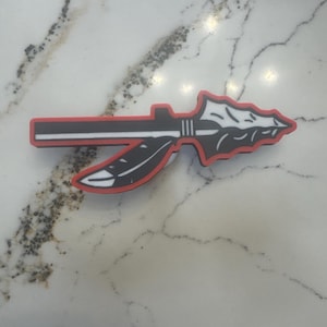 May include: A black and white arrowhead design with a red outline and a feather accent. The arrowhead is a symbol of Native American culture and is often used in jewelry and other crafts.
