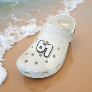 May include: White clog-style shoe with the number "61" in black and white on the side. The shoe has multiple ventilation holes and a textured design around the edges. The background shows a sandy beach and ocean waves.