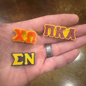May include: Three small, 3D printed Greek letter sorority pins. The pins are yellow and red with black outlines. The pins spell out "XO", "PIKA", and "&Sigma;N".