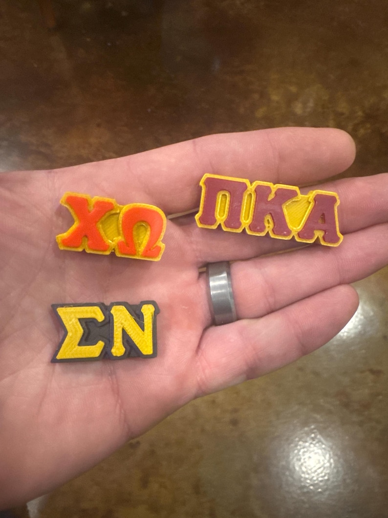May include: Three small, 3D printed Greek letter sorority pins. The pins are yellow and red with black outlines. The pins spell out "XO", "PIKA", and "&Sigma;N".
