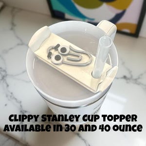 May include: White Stanley cup with a Clippy paperclip topper and a clear straw. The topper has a cartoon face design. The text on the cup reads "Clippy Stanley Cup Topper Available in 30 and 40 ounce."