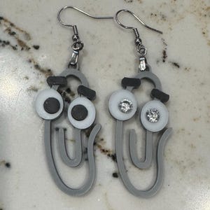 May include: A pair of gray paperclip earrings with white and black eyes. The eyes have a rhinestone in the center.