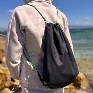 Könnte beinhalten: Ein schwarzer Kordelzug-Rucksack mit dunkelgrünen Kordelträgern. Die Tasche besteht aus einem einfarbigen schwarzen Stoff und wird über den Schultern getragen, wobei die grünen Kordeln die obere Öffnung zuziehen und bis zu den unteren Ecken reichen.