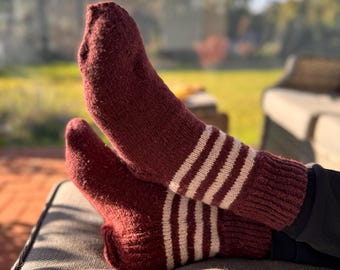 May include: A pair of burgundy socks with three white stripes on the upper ankle. The socks are thick and appear to be knitted, with a ribbed cuff. The socks are resting on a gray cushion outdoors.