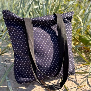 Op de afbeelding: Een marineblauwe draagtas met een wit hartjespatroon. De tas heeft zwarte hengsels en een ritssluiting. De tas ligt op zand, met groen gras op de achtergrond.