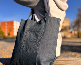 May include: A dark blue denim tote bag featuring a single exterior patch pocket with decorative stitching. The bag has two wide shoulder straps and is constructed from a durable fabric with visible seams, providing a structured rectangular shape.