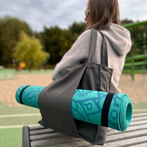 Puede incluir: Una esterilla de yoga turquesa enrollada con un dise&ntilde;o floral azul se guarda en un bolso tote gris con una correa negra. La bolsa descansa sobre un banco de madera. El fondo est&aacute; borroso, mostrando un parque.