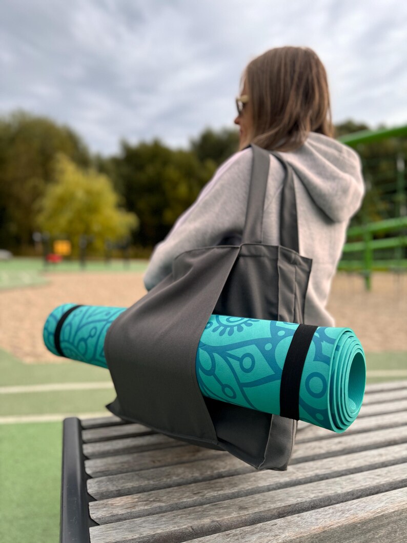 Puede incluir: Una esterilla de yoga turquesa enrollada con un dise&ntilde;o floral azul se guarda en un bolso tote gris con una correa negra. La bolsa descansa sobre un banco de madera. El fondo est&aacute; borroso, mostrando un parque.