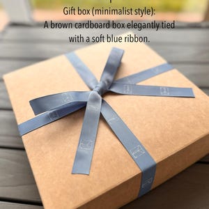 Puede incluir: Una caja de regalo de cart&oacute;n marr&oacute;n atada con una cinta azul claro. La caja es rectangular y de estilo minimalista. La cinta tiene un dise&ntilde;o sutil.