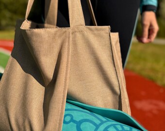 Puede incluir: Una bolsa de lona beige con correas largas sostiene una esterilla de yoga turquesa enrollada. La esterilla presenta un diseño de mandala turquesa. La bolsa la lleva una persona que lleva una chaqueta turquesa y leggings negros.