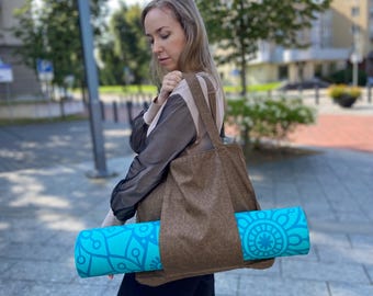 Puede incluir: Un bolso tote marrón con una esterilla de yoga turquesa enrollada. La esterilla de yoga tiene un patrón geométrico. El bolso se lleva al hombro. El fondo muestra una calle de la ciudad con árboles y edificios.