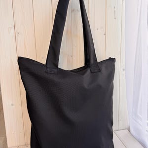 May include: A solid black tote bag featuring a rectangular body and two long shoulder straps. The bag is constructed from a smooth, opaque fabric and includes a zipper closure along the top edge, providing a clean and functional design for carrying items.