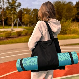 Puede incluir: Un bolso tote negro que lleva una esterilla de yoga enrollada. La esterilla de yoga es de color turquesa y azul con un dise&ntilde;o decorativo. La persona lleva una sudadera con capucha gris y un pantal&oacute;n de ch&aacute;ndal, de pie sobre una pista roja y verde.