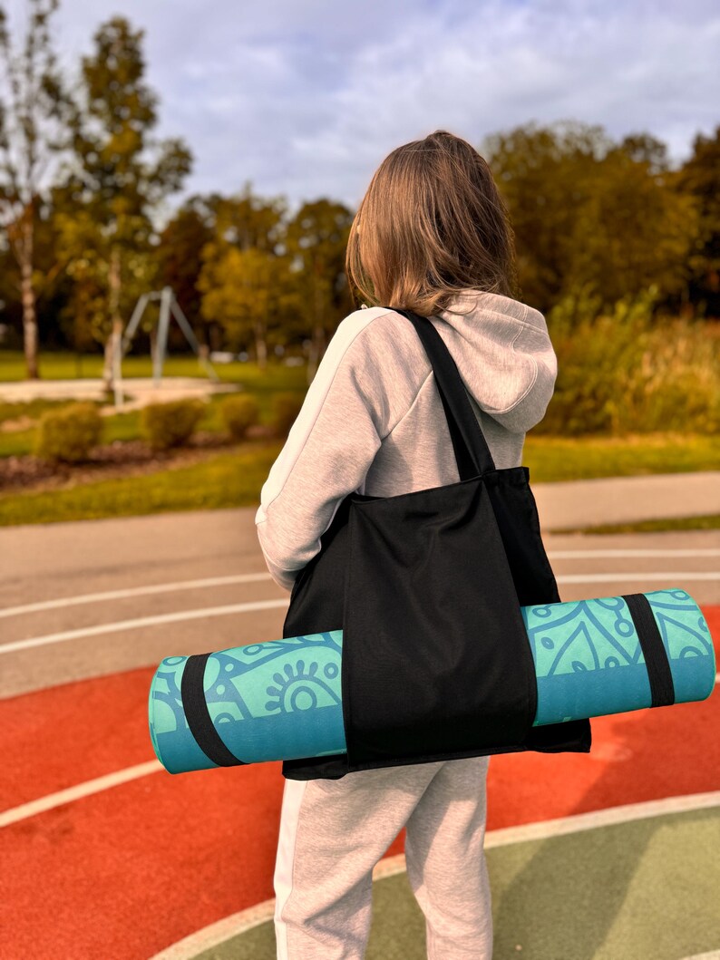 Puede incluir: Un bolso tote negro que lleva una esterilla de yoga enrollada. La esterilla de yoga es de color turquesa y azul con un dise&ntilde;o decorativo. La persona lleva una sudadera con capucha gris y un pantal&oacute;n de ch&aacute;ndal, de pie sobre una pista roja y verde.