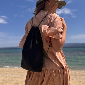 Può includere: Una persona indossa un vestito color pesca e un cappello di paglia su una spiaggia sabbiosa. Uno zaino nero con cordoncini verdi è indossato sulla spalla. Lo sfondo presenta un cielo blu e l'oceano.