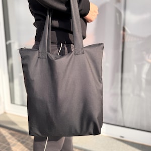 Può includere: Una borsa tote nera tinta unita con manici robusti. La borsa è rettangolare con chiusura a zip sulla parte superiore. La borsa è realizzata in un materiale liscio, forse nylon. La borsa tote è tenuta da una persona.