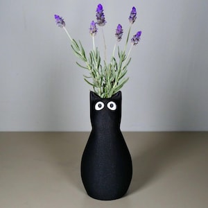 May include: A black cat-shaped vase with white eyes holds lavender sprigs with purple flowers and green stems. The vase is on a neutral-toned surface, with a plain gray background. The vase is a decorative item for the home.