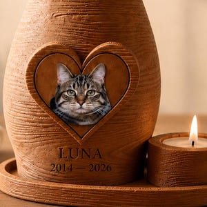 Puede incluir: Una urna conmemorativa con forma de gato y textura de madera marrón, con un recorte en forma de corazón que muestra la foto de un gato, el nombre LUNA y las fechas 2014-2026. Se asienta sobre una base a juego junto a un portavelas redondo encendido.
