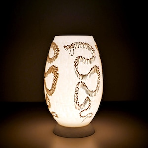May include: White ceramic lamp with a snake design etched into the surface. The lamp is lit and casting a warm glow.