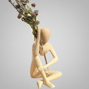 Op de afbeelding: Een beige sculptuur van een zittende figuur die een boeket gedroogde bloemen vasthoudt. De bloemen zijn bruin, paars en roze. De tekst "Arizona Hills" staat onderaan de afbeelding.