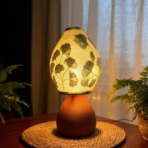 Puede incluir: Una lámpara de mesa decorativa con un brillo cálido. La lámpara presenta una base de terracota y una pantalla estampada con un diseño botánico. La lámpara está sobre una estera tejida.