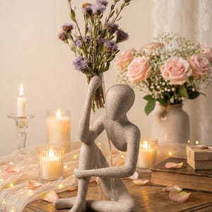 Könnte beinhalten: Eine graue Skulptur einer sitzenden menschlichen Figur, die eine Vase mit lila Blumen hält. Die Skulptur steht auf einem Holztisch mit Kerzen, Rosenblättern und einem Strauß rosa Rosen. Ein dekoratives, romantisches Thema.