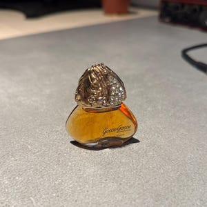 May include: A small, clear glass bottle of Gucci Gucci by Napoleon perfume. The bottle has a gold cap with rhinestones and the words "Gucci Gucci by Napoleon Toilette" are printed on the bottle.
