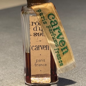 Puede incluir: Un pequeño frasco de perfume Carven de vidrio transparente con una tapa dorada y una etiqueta amarilla con texto verde que dice "Carven Paris parfums France" y "ro d'un soir". El frasco también tiene una etiqueta que dice "carven paris france" y "net cont."