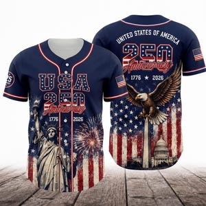 May include: A navy blue baseball jersey featuring a front graphic of the Statue of Liberty, fireworks, and the text USA 250 Anniversary 1776 2026. The back displays a bald eagle, the Washington Monument, the Capitol building, and the text United States of America.