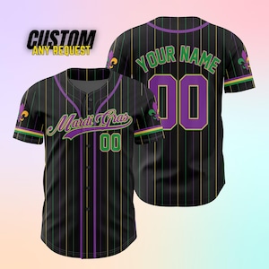 K&ouml;nnte beinhalten: Schwarzes und gr&uuml;nes Baseballtrikot mit lila Streifen und einem Mardi Gras-Design. Das Trikot hat eine Fleur-de-lis am linken &Auml;rmel und die Nummer 00 auf der Vorderseite. Die R&uuml;ckseite des Trikots tr&auml;gt den Text "YOUR NAME" und die Nummer 00.