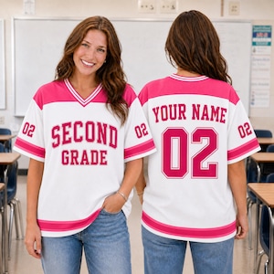 May include: A white and pink V-neck jersey featuring the text SECOND GRADE on the front and the text YOUR NAME with the number 02 on the back. The sleeves and hem have pink and white stripes, and the number 02 is printed on both sleeves.