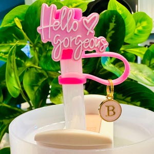 May include: A pink and white water bottle accessory with the words "Hello gorgeous" in a script font. The accessory has a pink loop and a gold charm with the letter "B" on a pink background. The accessory is set against a background of green leaves.