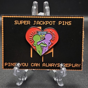 May include: A black card with orange text that reads "Super Jackpot Pins" and a red heart with two figures inside. The figures are green and purple and are holding hands in front of a pinball machine. The text at the bottom of the card reads "Pins you can always replay."
