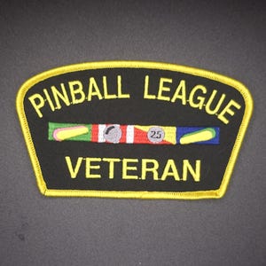 May include: A black embroidered patch with the words "PINBALL LEAGUE VETERAN" in yellow. The patch has a yellow border and a colorful pinball design in the center, including the number "25".