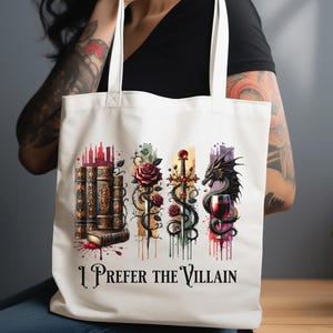Pode incluir: Sacola de tecido branca com um design colorido que apresenta uma pilha de livros, uma espada, uma rosa, um drag&atilde;o e uma ta&ccedil;a de vinho. O texto "I Prefer the Villain" est&aacute; impresso na sacola.