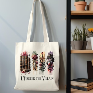 Pode incluir: Uma bolsa tote de lona branca com uma ilustra&ccedil;&atilde;o colorida de livros, uma rosa, uma espada, um drag&atilde;o e uma ta&ccedil;a de vinho. O texto "I Prefer the Villain" est&aacute; impresso na bolsa.